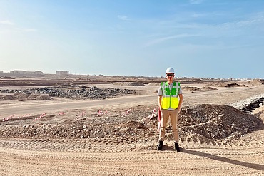 A man in a white construction site helmet and high-visibility vest stands with his hands in his pockets in front of a large construction site in Oman. Sandy earth below, blue sky above.