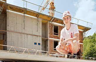 Foto: Ein Lehrling sitzt in der Hocke auf einem Rohbauelement und blickt in die Kamera