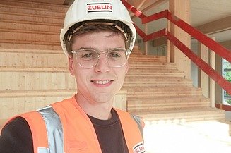 Selfie eines jungen Mannes mit Bauhelm, im Hintergrund ist eine Treppe aus Holz zu sehen