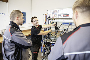 Photo Three men stand in front of a workbench, the one behind explains