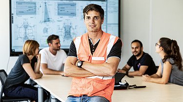 Colleagues talking in the background - one colleague sits in focus and looks into the camera