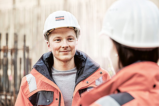 Foto Mitarbeiter mit Bauhelm blickt Mitarbeiterin mit Bauhelm an