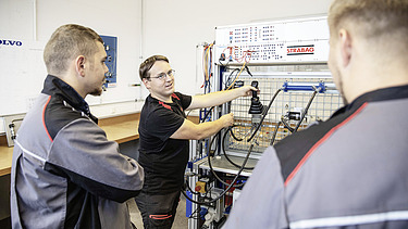 Photo Three men stand in front of a workbench, the one behind explains